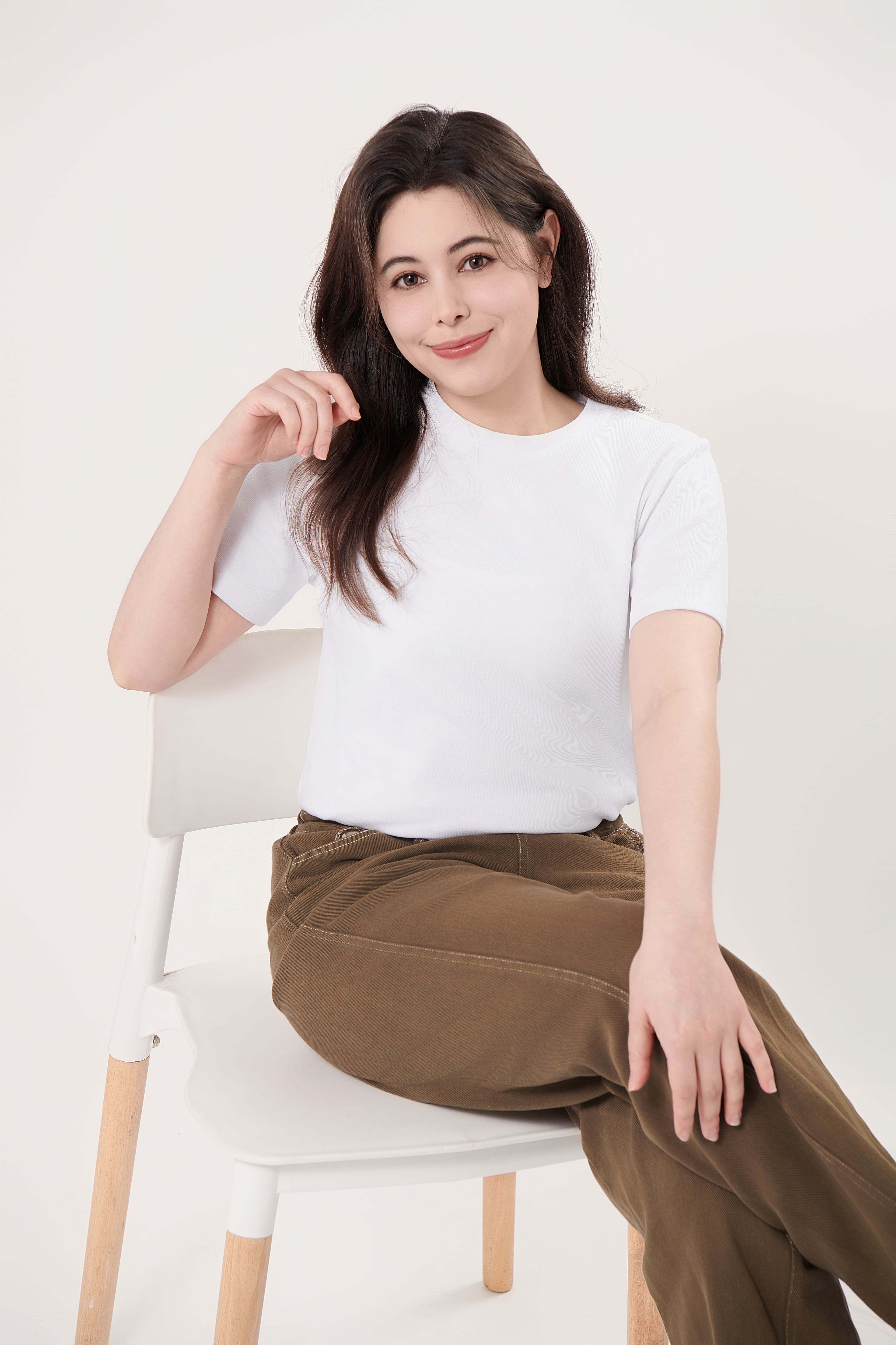 Smiling woman wearing a white t-shirt and brown pants sitting on a white chair.